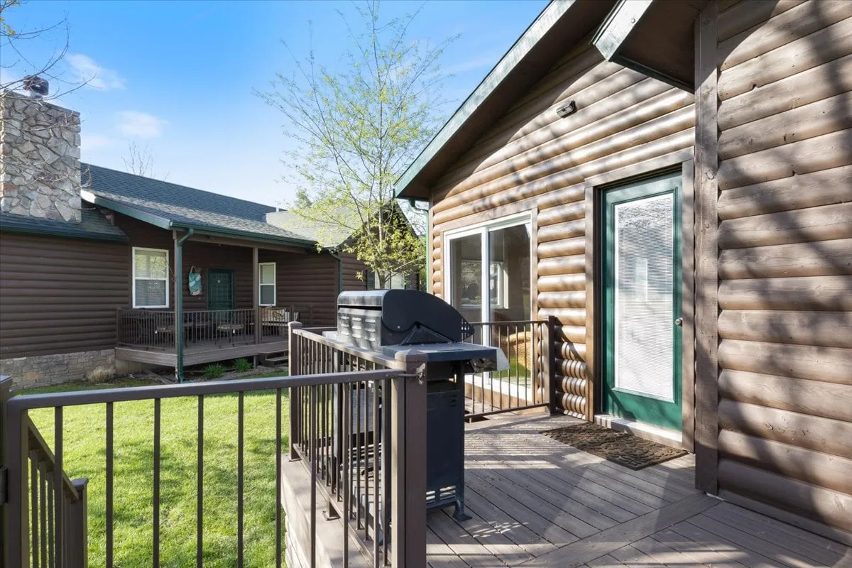 An inviting exterior deck with a grill, deck railing, log cabin siding, and a green door. The setup is ideal for outdoor dining, barbecuing, and soaking in the outdoors, with cozy touches that encourage relaxation and socializing.
