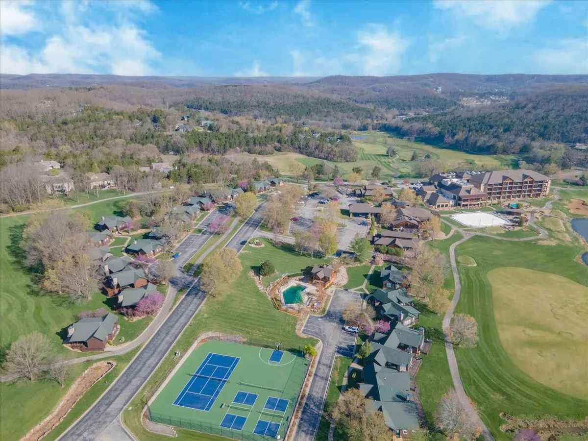 This image features the tennis courts and a swimming pool visible in the foreground, with a hotel building and wooden cabins nestled among landscaped gardens behind. The scene conveys leisurely activities such as tennis, swimming, and relaxing in a communal resort setting, all under pleasant natural light.