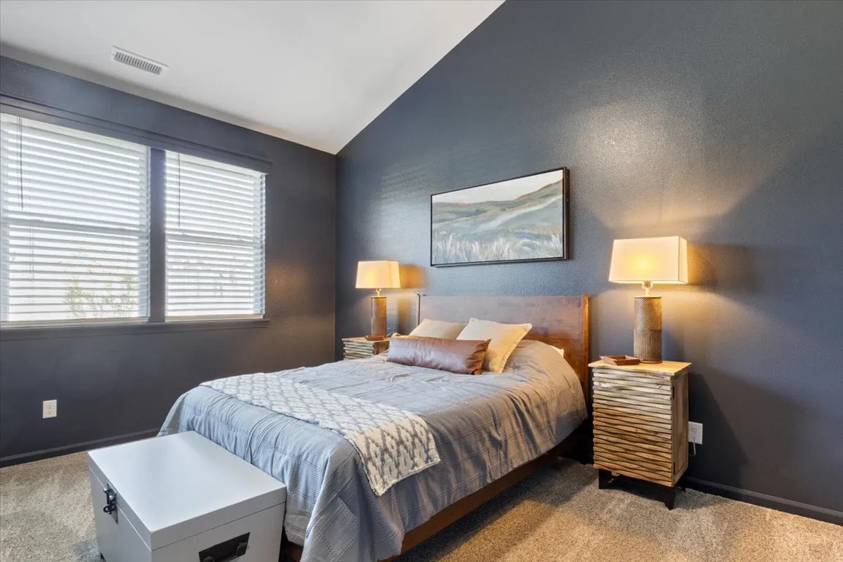 A serene bedroom with a queen bed, decorative pillows, nightstands with lamps, and large windows with blinds. The space is bathed in soft natural light, highlighting comfortable bedding and tranquil design—ideal for a relaxing retreat.