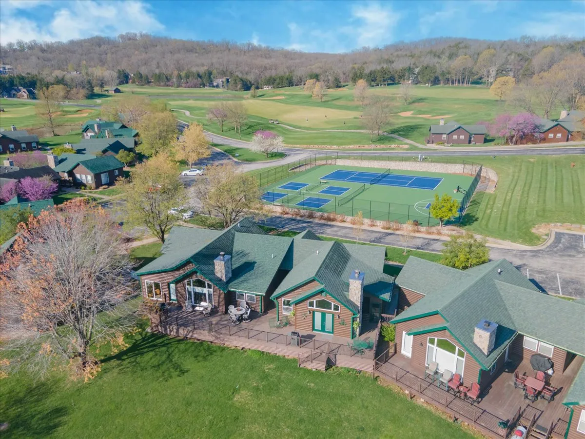 The image showcases two charming cottages with inviting decks overlooking a pristine golf course, bordered by tennis courts. The sun is setting, casting a warm glow over the green rolling fairways and outdoor spaces, suggesting both relaxation and recreation in a serene community setting.