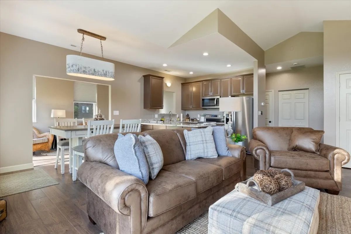 An open interior featuring a brown leather sofa adjacent to a dining table and kitchen island. The space is accented by a decorative light fixture, blending living, dining, and kitchen areas in a cohesive, open-concept layout. Warm tones and inviting furnishings encourage togetherness and conversation.