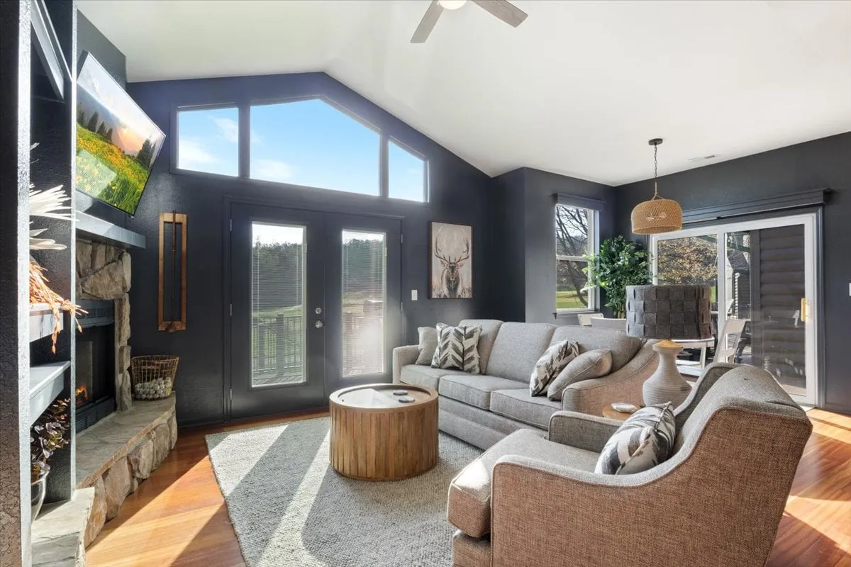 A comfortable living room with large windows offering scenic views, plush seating, a decorative fireplace, a large TV, and a stylish coffee table. The mood is tranquil and inviting, suited for relaxation or socializing.
