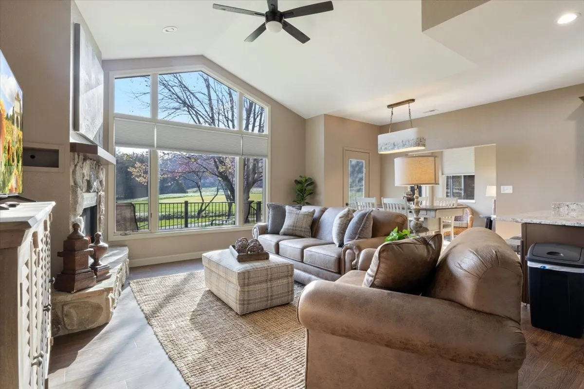 A spacious living room bathed in natural light from large windows, featuring a comfortable seating arrangement centered around a decorative stone fireplace and an entertainment center with TV. The space invites groups to gather for conversation, games, or relaxation after a day of adventure.