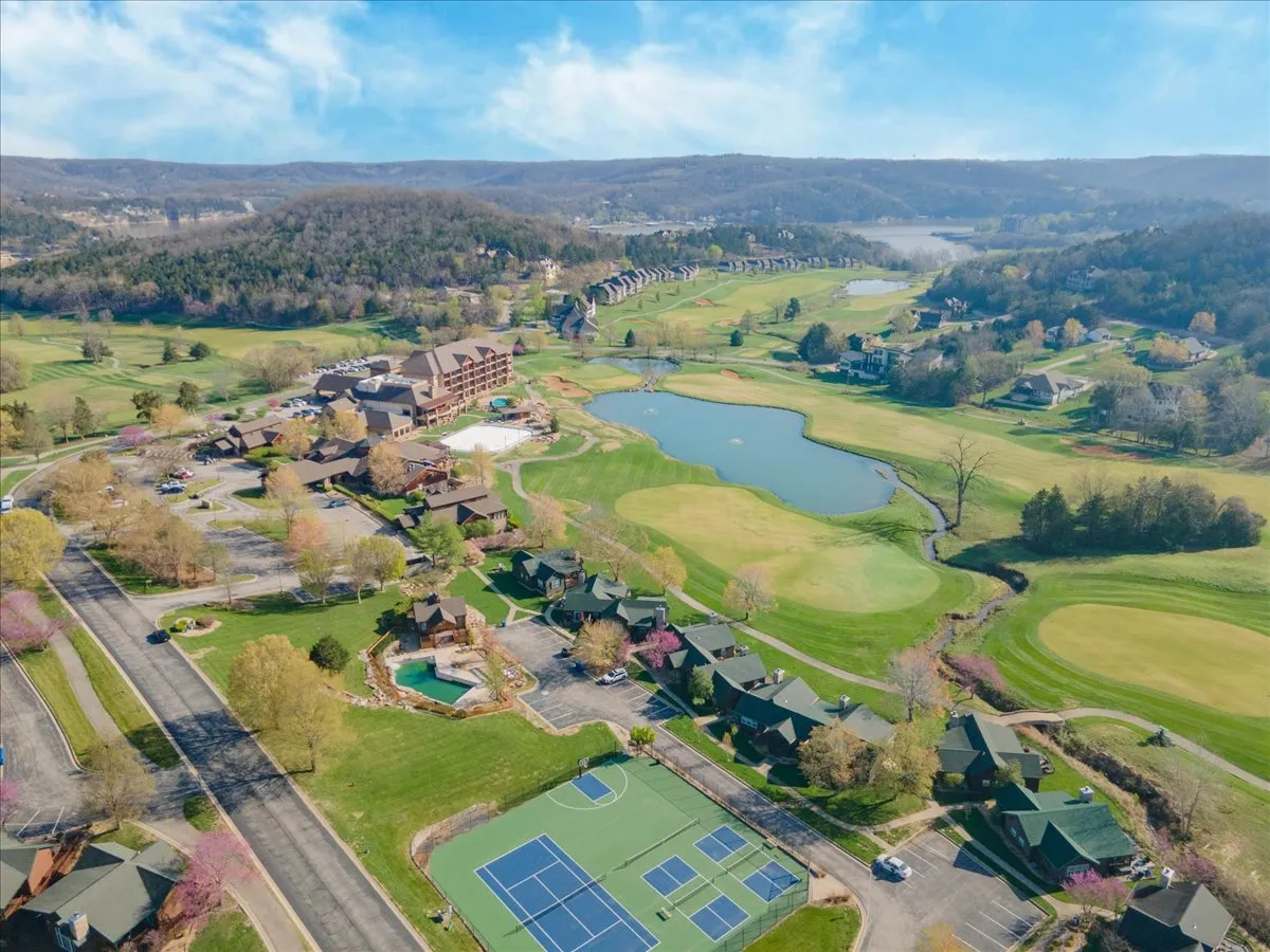 An exterior overview emphasizing the resort building set beside a shimmering lake and expansive golf course, with visible tennis courts and mature trees framing the scene. The mood is both recreational and tranquil.